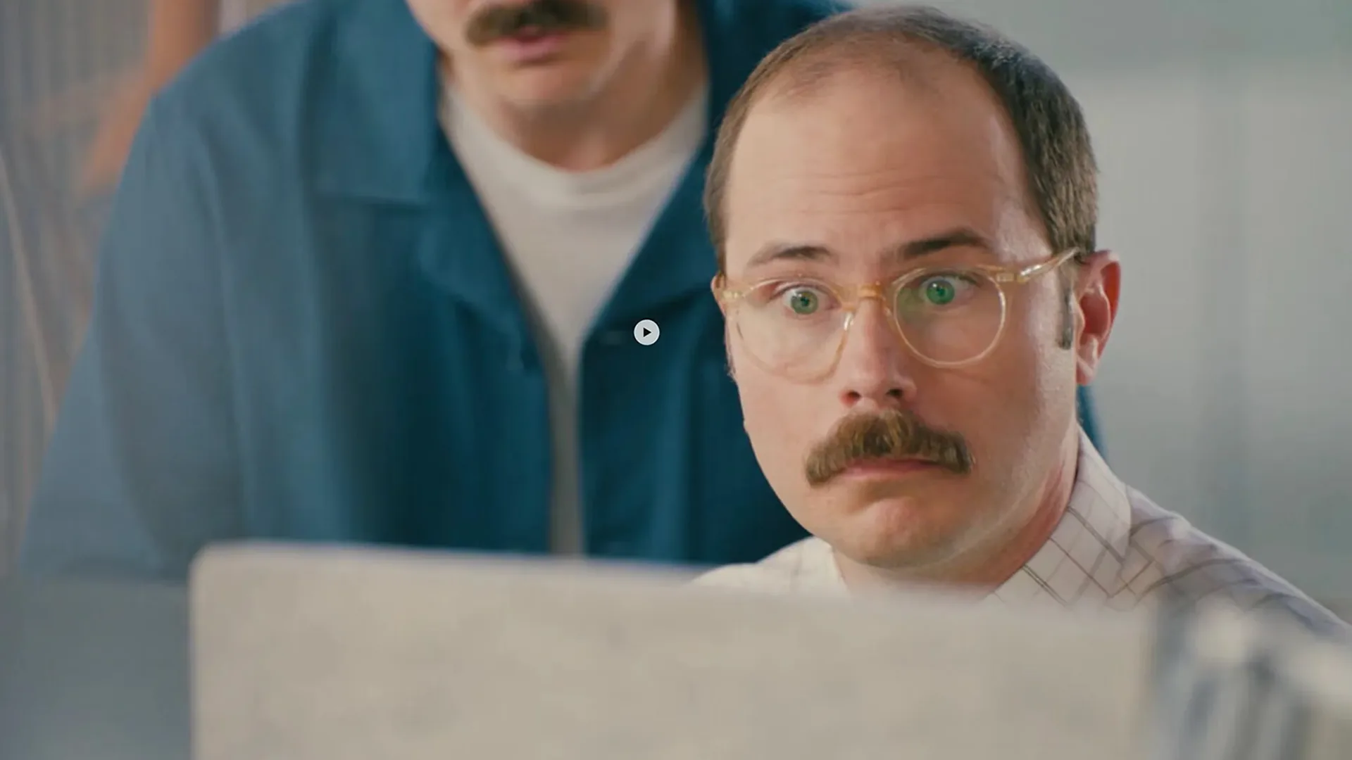 Close-up of a wide-eyed, mustached man in glasses staring at a laptop screen while another man leans in over his shoulder, both reacting to something on the display