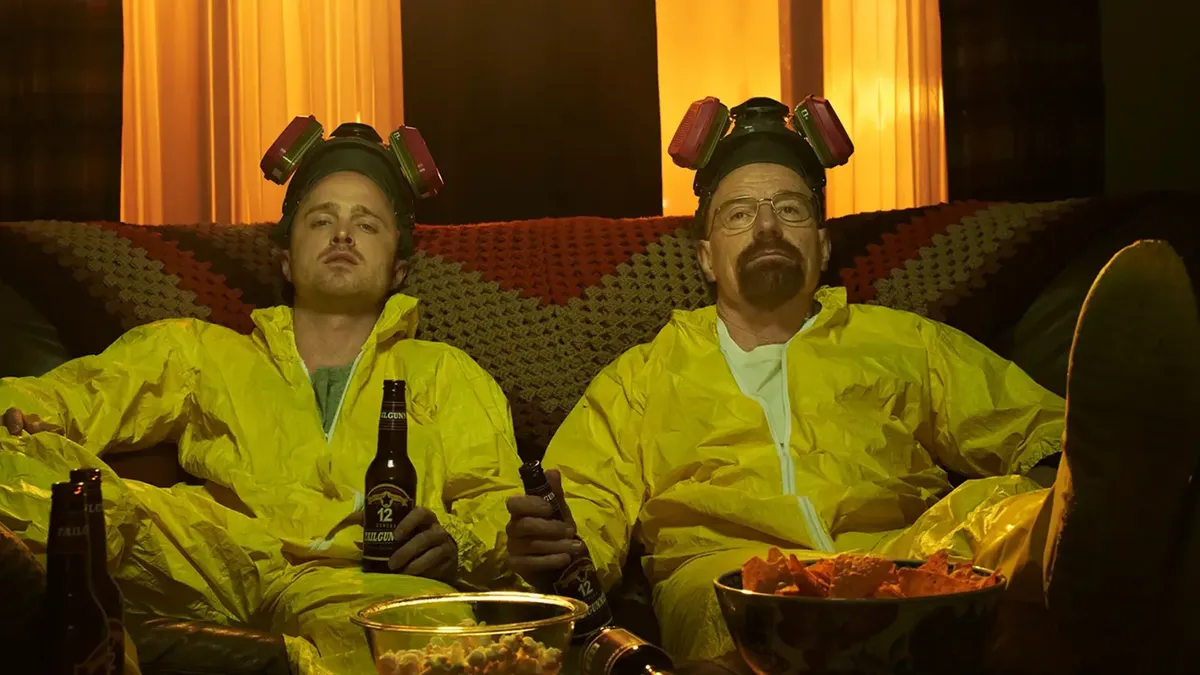 Two collaborators in matching yellow hazmat suits and respirators slumped together on a couch with beers and snacks after intense shared work, evoking a tight two-person crew