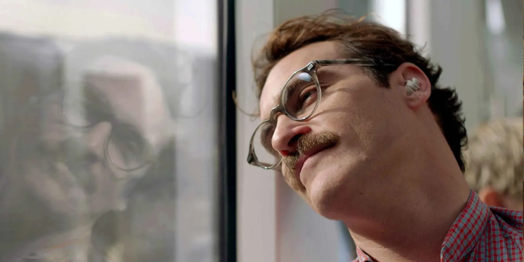 Joaquin Phoenix as Theodore in the film Her, wearing round glasses and an earpiece, gazing out a window with a soft smile as he talks to an invisible AI